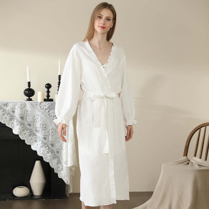 Woman wearing white cotton lace kimono robe standing in cozy home setting with candles and lace tablecloth in the background.